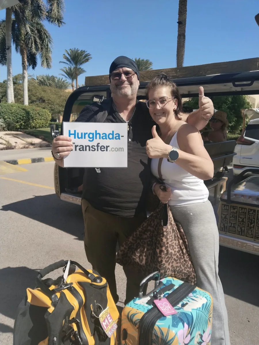 Two smiling guests with their luggage and a Hurghada-Transfer.com sign outside their Red Sea hotel