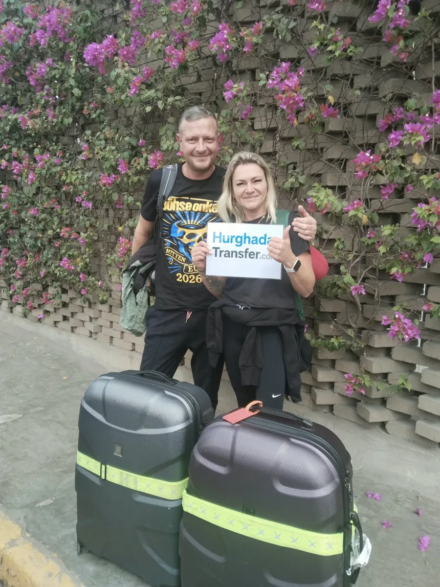A couple with two large suitcases holding a Hurghada-Transfer.com sign in front of a wall of bougainvillea