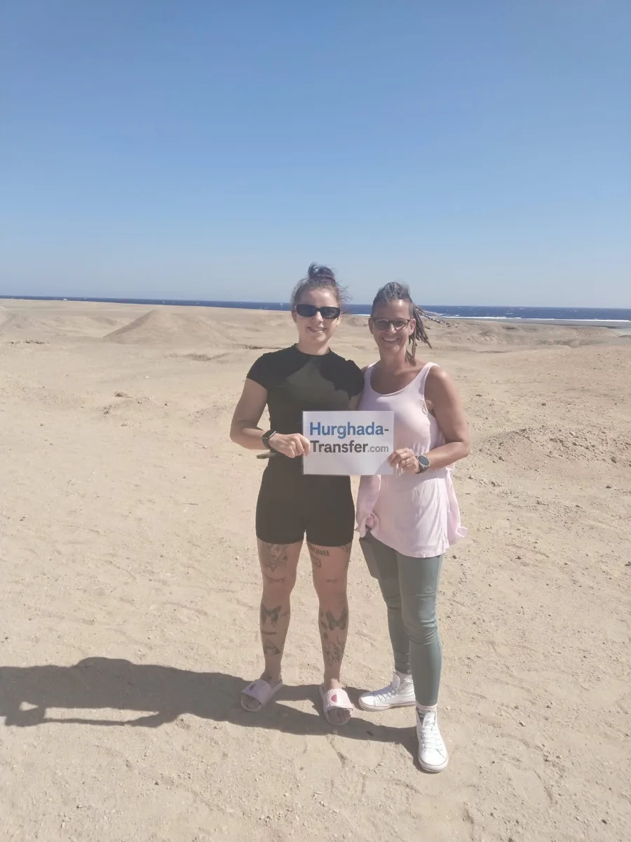 Two travellers holding a Hurghada-Transfer.com sign on a sandy stretch of the Red Sea coast